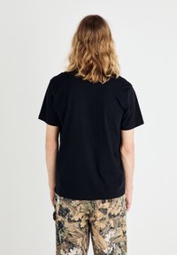 T-shirt in cotone nero con colletto rotondo classico e maniche corte, vista da dietro. Abbinata a pantaloni cargo con fantasia.