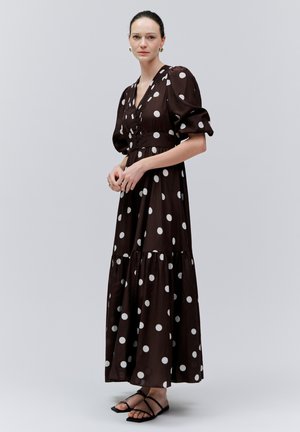 Femeie în picioare purtând o rochie lungă neagră cu buline albe, mâneci bufante și sandale negre cu barete, pe un fundal simplu, deschis la culoare.