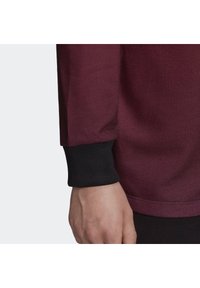 Felpa bordeaux con tessuto testurizzato e polsini neri a costine, che presenta un design delle maniche aderente e una sottile rifinitura del bordo.