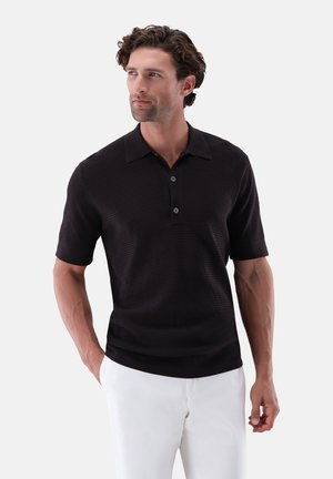 Mann mit lockigem Haar, der ein schwarzes kurzärmeliges strukturiertes Poloshirt und weiße Hose trägt, steht mit einer Hand in der Tasche.