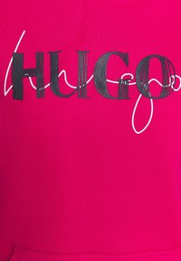 Felroze stof met een geribbelde textuur, met het woord "HUGO" in dikke zwarte letters en witte accenten. Eenvoudig ontwerp, zonder extra details.