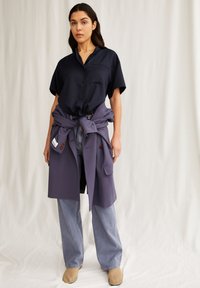 Marineblause mit kurzen Ärmeln, lila Jacke um die Taille gebunden, weit geschnittene hellblaue Jeans und beigen Schuhen. Weicher Stoff mit entspanntem Design.