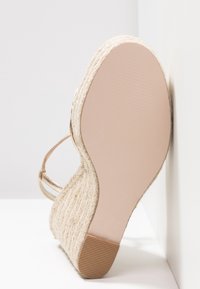Sandały na koturnie z plecionym obcasem z juty, w jasnym beżowym kolorze, gładka gumowa podeszwa oraz cienkie metaliczne paski. Szczegółowa tekstura na obcasie.