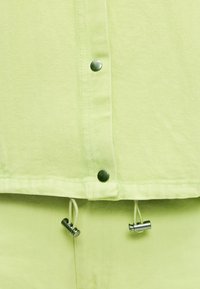 Ανοιχτό πράσινο ύφασμα με μεταλλικά κουμπιά τύπου snap και κορδόνι με ασημένιες αντικλείδες στο κάτω άκρο.