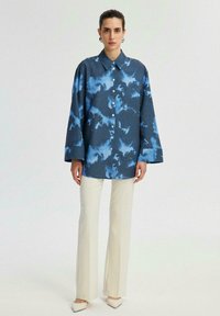 Camicia abbottonata di colore blu scuro con un motivo di nuvole azzurro chiaro, caratterizzata da maniche lunghe, colletto e vestibilità ampia. Abbinata a pantaloni color crema.