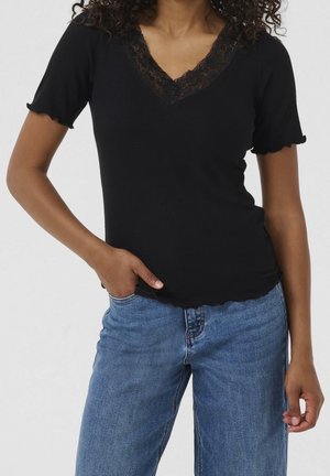 Kvinde iført sort kortærmet V-hals top med blondekant og blå denimjeans, med den ene hånd i lommen mod en ensfarvet baggrund.