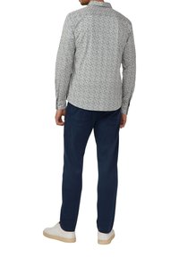 Man in een gestreept overhemd met lange mouwen, marineblauwe broek en witte sneakers, staand met zijn rug naar de kijker op een witte achtergrond.