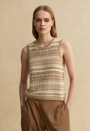 Giovane donna che indossa un top lavorato a maglia senza maniche beige e marrone con motivi e pantaloni marroni a vita alta, in piedi davanti a uno sfondo beige uniforme.