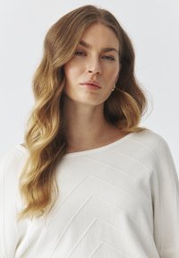 Hvid sweater med et tekstureret chevronmønster. Fremstillet af blødt stof, med en bred halsudskæring og en afslappet pasform. Langt, bølget hår, der rammer ansigtet.