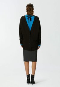 Tamno smeđi kardigan s dubokim V-izrezom, s redom dugmadi, složen preko plave košulje s crnim trakama. Siva suknja i crne čizme.