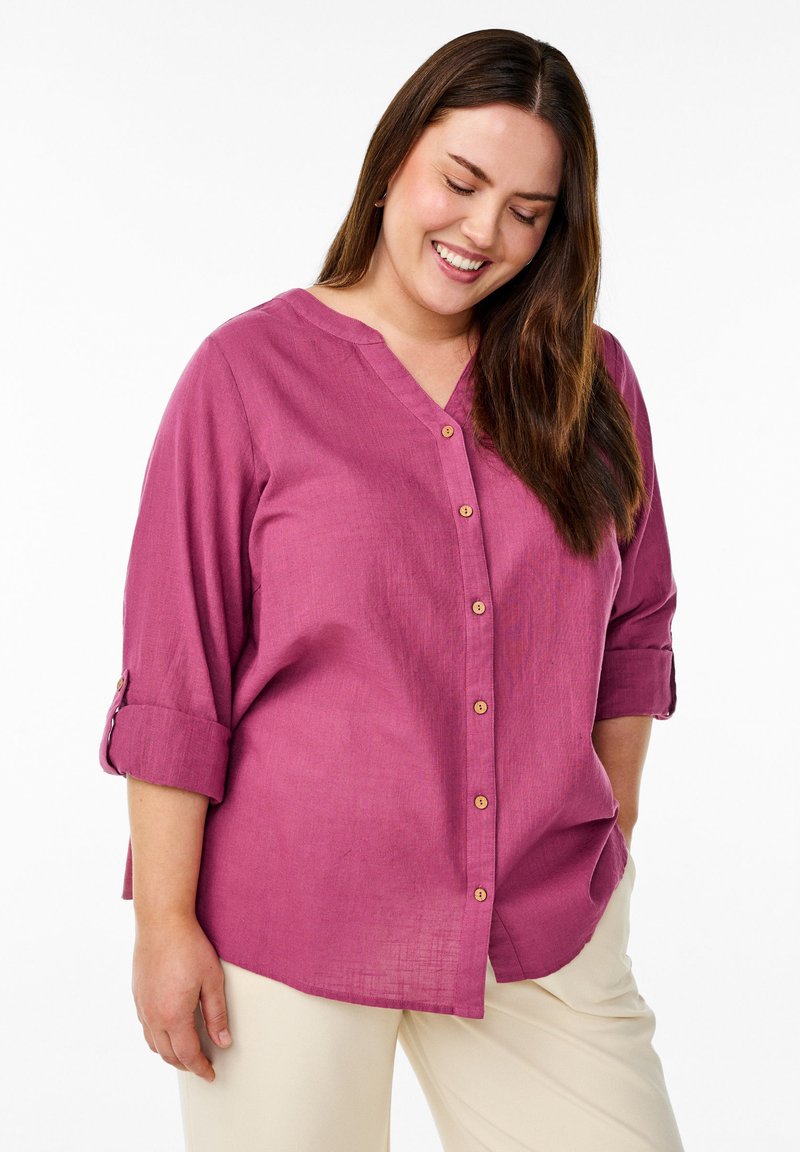 Camicia fucsia con colletto a V, maniche arrotolabili e bottoni in legno. Indossata con pantaloni chiari, che mostrano una vestibilità rilassata.
