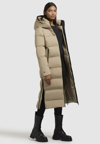 Piumino beige con cappuccio, lunghezza lunga e sezioni trapuntate. Presenta cerniere nere e foderato interno, abbinato a stivali neri con lacci.