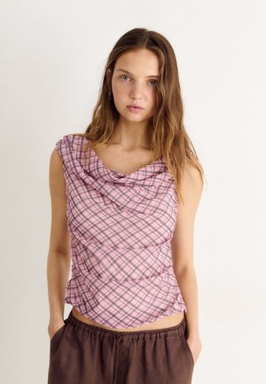 Jeune femme aux longs cheveux bruns portant un haut sans manches à carreaux roses et un pantalon marron, debout devant un fond blanc uni.