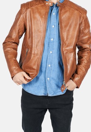 Personne portant une veste en cuir beige par-dessus une chemise bleue à boutons et un pantalon noir, ajustant la fermeture éclair de la veste avec les deux mains.