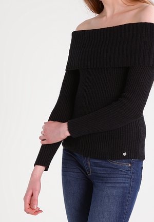 Femme portant un pull côtelé noir à épaules dénudées et un jean bleu, la main gauche tenant son avant-bras droit.