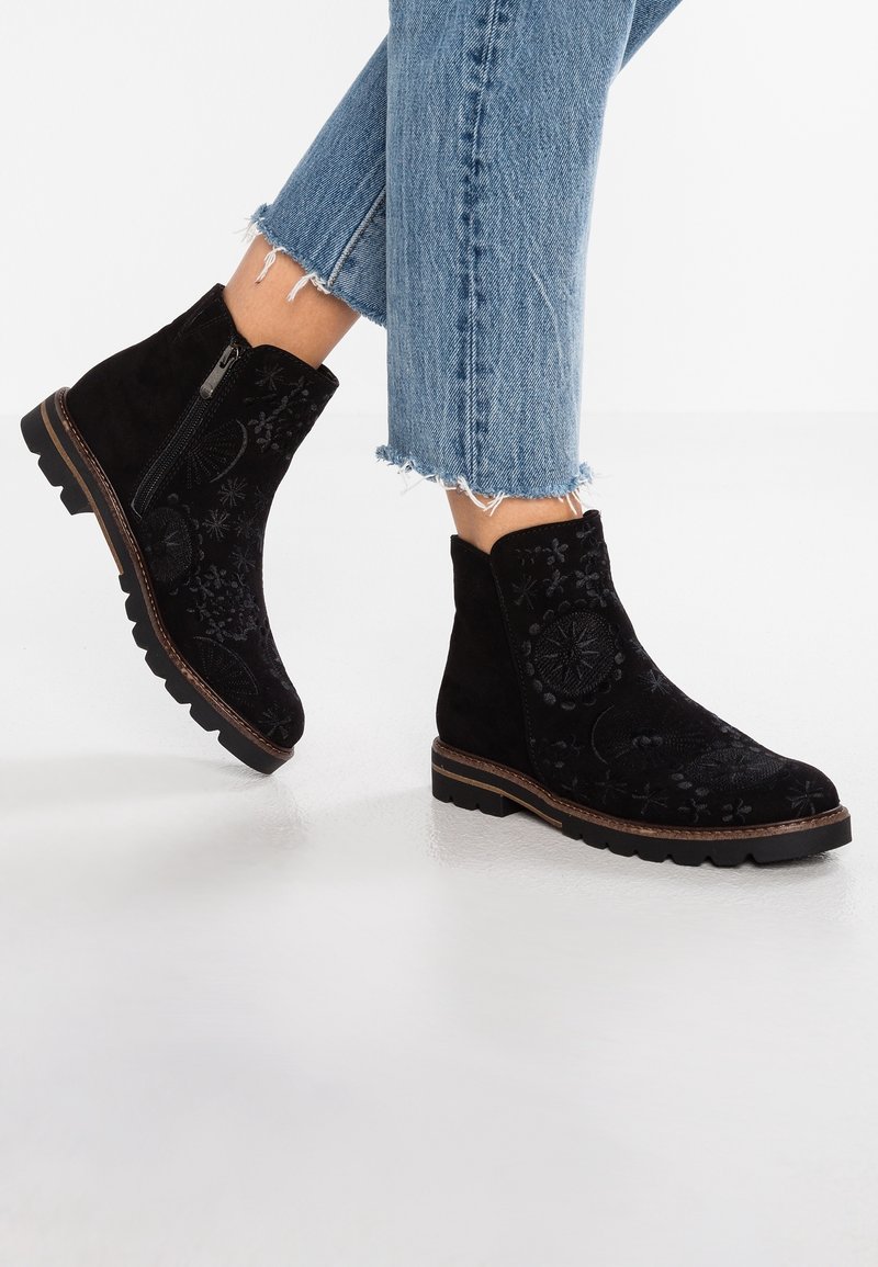 Bottines noires à la cheville avec broderie florale et fermetures éclair latérales, portées avec un jean bleu coupé effiloché sur fond blanc.