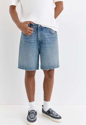 Personne portant un short en jean bleu délavé de longueur moyenne, un t-shirt blanc, des chaussettes blanches et des baskets noires à enfiler avec une broderie blanche.
