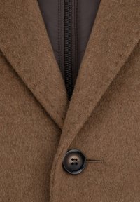 Brązo wełniany płaszcz z klapami, z przednim zamkiem błyskawicznym i pojedynczym czarnym guzikiem po lewej stronie. Miękka faktura.