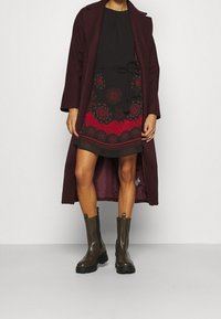 Hnědočervený dlouhý kabát přes černé šaty s červenými květinovými vzory a zavazováním v pase. Hnědé kotníkové boty s robustní podrážkou. Hladké textury po celém outfitu.