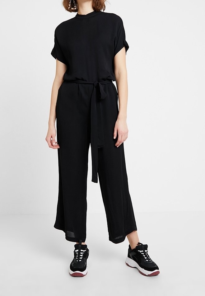 Femme portant une combinaison noire à manches courtes avec un lien à la taille et des baskets épaisses noires et blanches, debout sur un sol blanc simple.