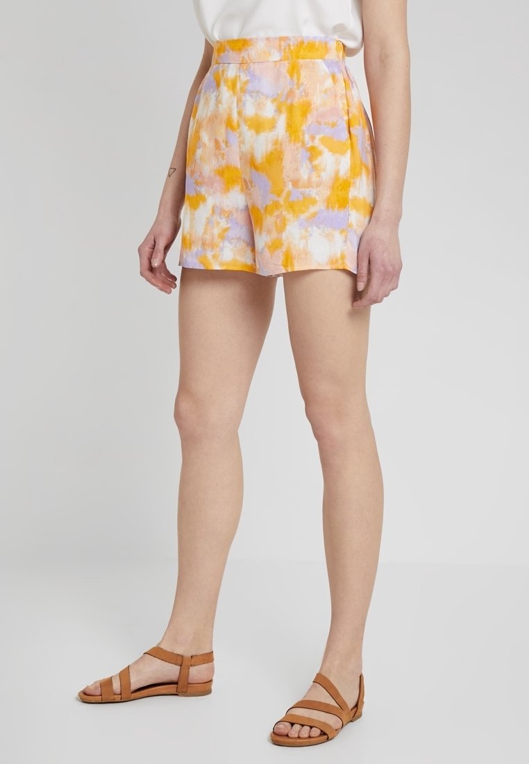 Femme portant un short tie-dye orange, lavande et blanc, associé à des sandales plates couleur beige, debout devant un fond clair uni.