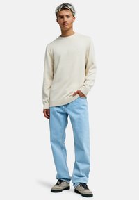 Nuori mies seisoo valkoisella taustalla, päällään kermaisen värinen neulepaita, vaaleansiniset farkut ja beiget tennarit, käsi taskussa.