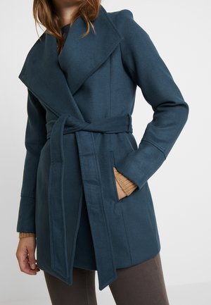 Kobieta ubrana w zielonkawo-niebieski wełniany płaszcz z paskiem i szerokimi klapami, z ręką w kieszeni, na tle prostego tła, z beżowym swetrem i brązowymi spodniami.