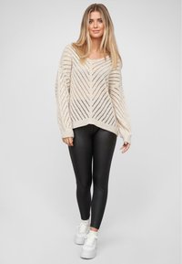 Cremfarbener Strickpullover mit offenem Chevronmuster, weiten Ärmeln und V-Ausschnitt, kombiniert mit schwarzen Leggings und weißen Sneakers.
