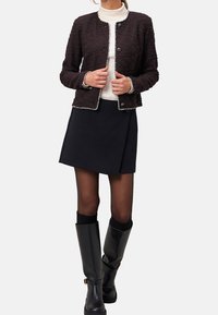 Veste noire texturée avec bordure argentée, portée sur un col roulé blanc. Associée à une jupe foncée et des bottes noires montantes jusqu'aux genoux.