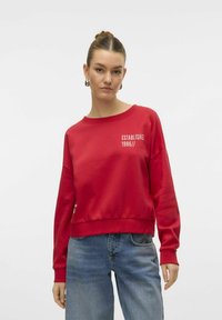 Sweatshirt rouge en matériau doux avec un col rond et des épaules tombantes. Présente un texte blanc, "ÉTABLI EN 1986//," sur le devant.