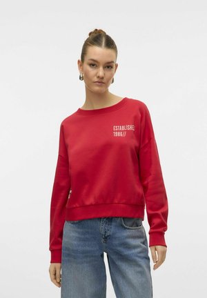 Sweatshirt rouge en matériau doux avec un col rond et des épaules tombantes. Présente un texte blanc, "ÉTABLI EN 1986//," sur le devant.