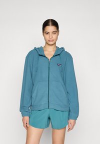 Blue zip-up hooded sweatshirt made of soft fabric, featuring a front pocket, drawstring hood, and logo patch on the left chest.