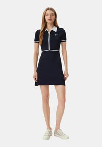 Laivaston sininen polo-mekko, jossa on lyhyet hihat, valkoinen kaulus ja kanttaus, puolivetoketju edessä, sileä ja vartalonmyötäinen muotoilu, yhdistettynä vaaleisiin tennareihin.