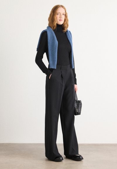 Haut noir à col roulé associé à un pantalon noir ample taille haute. Un gilet en tricot bleu clair repose sur les épaules. Sac à main noir.