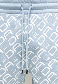 Light blue sweatpants featuring a white repeated logo pattern, drawstring waist, and black-tipped cord. Soft texture and relaxed fit.