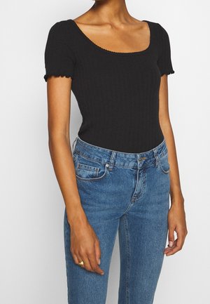 Frau trägt ein enganliegendes schwarzes kurzärmeliges, geripptes Oberteil mit Rundhalsausschnitt und blaue, hoch taillierte Jeans, steht vor einem schlichten Hintergrund.