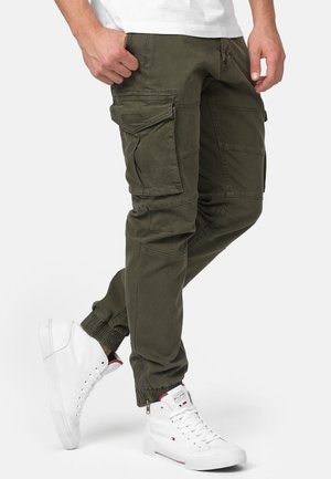 Homme portant un pantalon de jogging cargo vert olive avec des bas élastiques et des baskets hautes blanches, debout avec une jambe légèrement pliée.