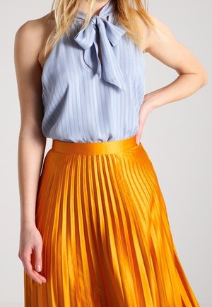 Woman wearing a light blue sleeveless blouse with a bow and a high-waisted, pleated bright orange skirt, posing with one hand on hip.