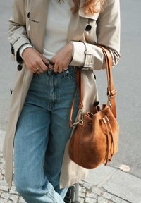 Tan suede bucket bag with drawstring closure, and adjustable strap. Worn with beige trench coat and light blue high-waisted jeans.