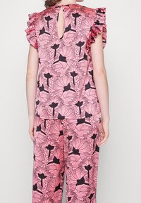 Růžový top s květinovým vzorem, řasenými rukávy a výřezem na zádech. Materiál se zdá být lehký a hladký. V kombinaci s odpovídajícími kalhotami.