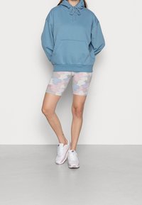 Felpa con cappuccio in cotone blu con coulisse, abbinata a pantaloncini da ciclismo mimetici multicolori e sneaker bianche con dettagli rosa.