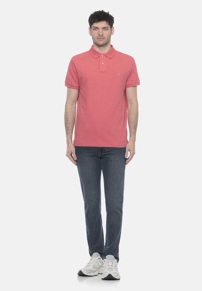 Homme debout portant un polo rose corail, un jean bleu foncé et des baskets blanches sur un fond blanc uni.