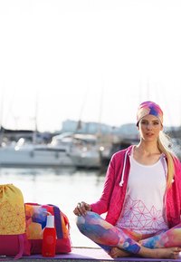 Frau in bunter Sportkleidung, die im Schneidersitz auf einem Steg meditiert, mit Booten im Hintergrund und Taschen neben sich.