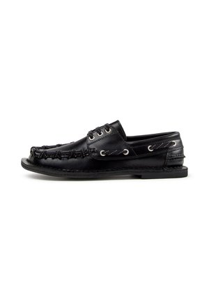 Chaussure style mocassin en cuir noir avec coutures visibles, détails de lacets autour de l’orteil et semelle en caoutchouc plate sur fond blanc.