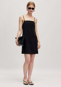Schwarzes Slipkleid mit dünnen Trägern, glatter Textur, knielang, kombiniert mit einer schwarzen Clutch und Flip-Flops, getragen vor einem neutralen Hintergrund.