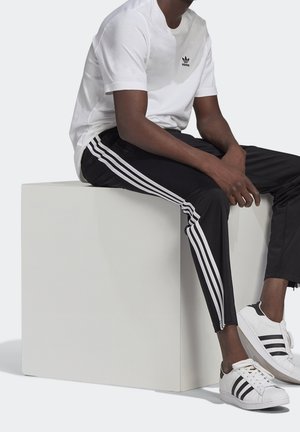 Man som bär vit Adidas t-shirt, svarta Adidas träningsbyxor med tre vita ränder och vita Adidas-sneakers, sittande på en vit kub.