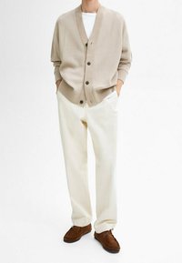 Beige Rippstrick-Cardigan mit fünf Knöpfen, getragen über einem weißen T-Shirt, kombiniert mit cremefarbenen, weiten Hosen und braunen Wildlederschuhen.