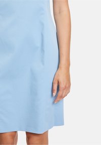 Jasnoniebieska sukienka z gładkiego materiału, o dopasowanym kroju i subtelnych szwach bocznych. Dłoń spoczywa przy dolnym brzegu, eksponując naturalne paznokcie.