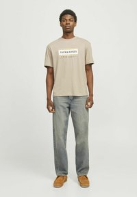 Beige Baumwoll-T-Shirt mit schwarzem und goldenem Logo-Graphic, kombiniert mit hellblauen Jeans und hellbraunen Wildlederschuhen. Flacher Schnitt und kurze Ärmel.
