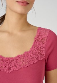 T-shirt à manches courtes en rose profond avec un détail de dentelle florale texturée au niveau du col, présentant un design à encolure dégagée large et un tissu côtelé.